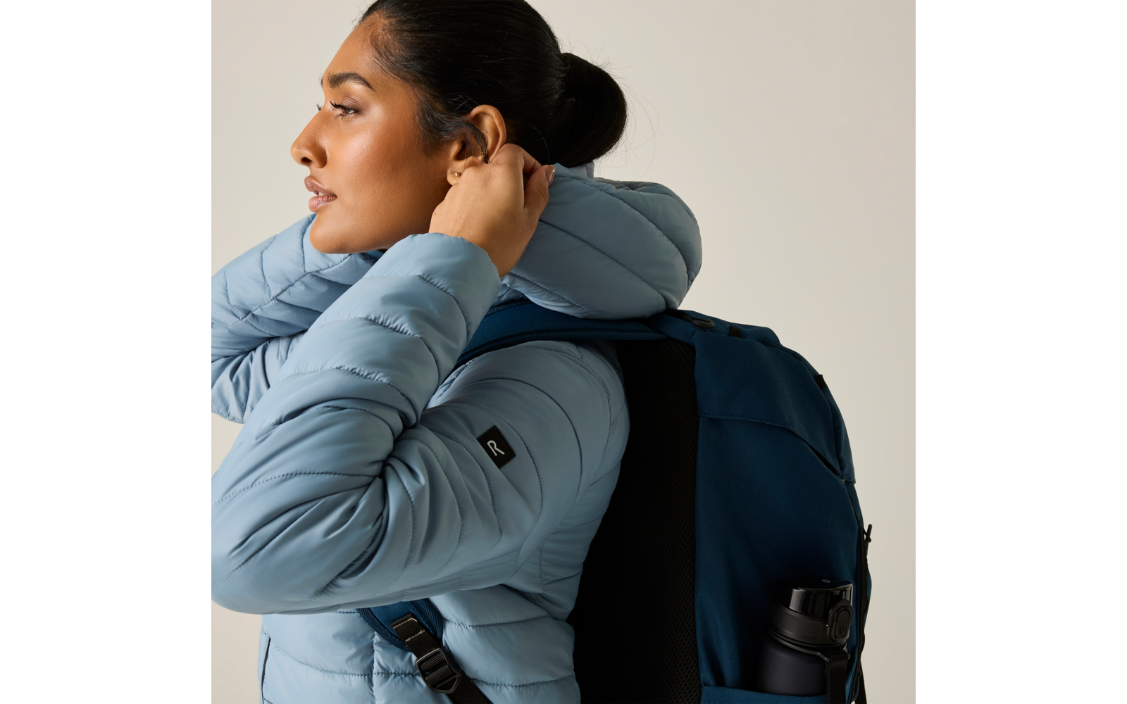 Person im Profil trägt hellblaue Steppjacke und dunkelblauen Rucksack, Hand am Ohr; dunkel gebundenes Haar, einfarbiger heller Hintergrund