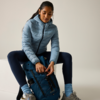Person sitzt auf weißem Block, trägt hellblaue Steppjacke, dunkle Leggings und graue Schuhe, öffnet einen blauen Rucksack