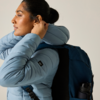 Person im Profil trägt hellblaue Steppjacke und dunkelblauen Rucksack, Hand am Ohr; dunkel gebundenes Haar, einfarbiger heller Hintergrund
