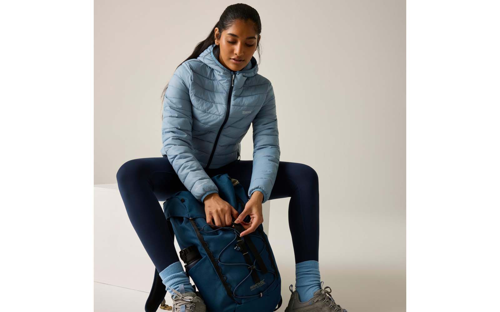 Person sitzt auf weißem Block, trägt hellblaue Steppjacke, dunkle Leggings und graue Schuhe, öffnet einen blauen Rucksack