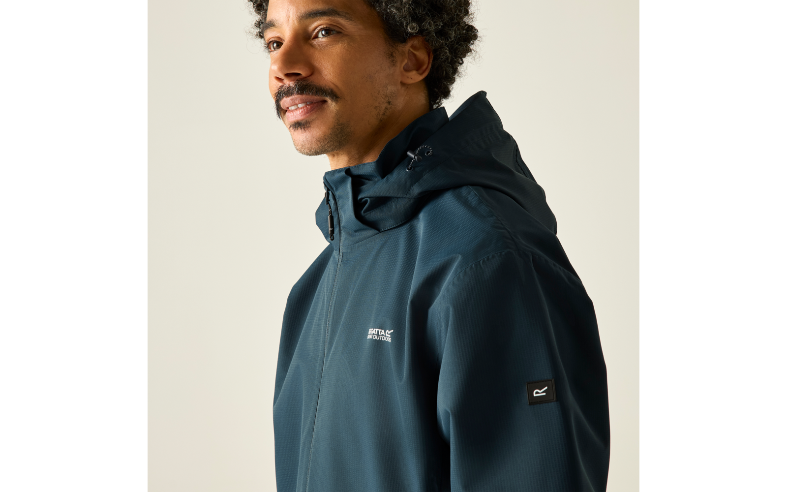 Person mit Schnurrbart und kurzen lockigen Haaren, trägt eine dunkelblaue Kapuzenjacke mit kleinem weißem Brustlogo und schwarzem Ärmellogo, vor hellem Hintergrund, Blick zur Seite