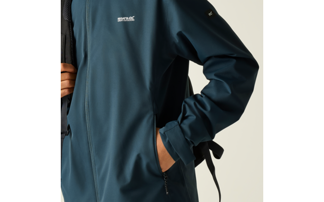 Nahaufnahme eines Oberkörpers in petrolfarbener Jacke, eine Hand in der Tasche, die andere hält einen Rucksackträger, kleines weißes Logo auf der Brust