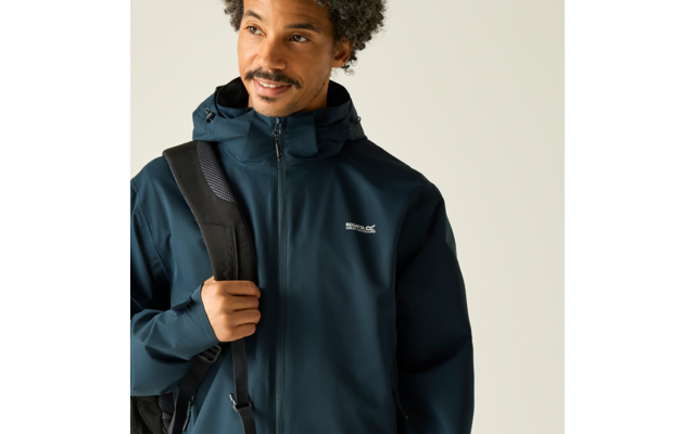 Person mit lockigem Haar und kurzem Bart, trägt dunkelblaue Kapuzenjacke mit geschlossenem Reißverschluss, Träger einer Tasche über der Schulter, Blick zur Seite vor hellem Hintergrund