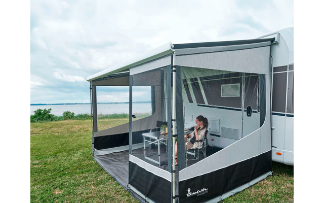 Personne assise à une table dans une bâche fermée attachée à une caravane, caravane à droite, herbe et lac en arrière-plan, ciel nuageux