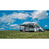 Camping-car blanc avec auvent déployé sur un champ herbeux, deux personnes assises dessous sur des chaises de camping, ciel bleu avec nuages