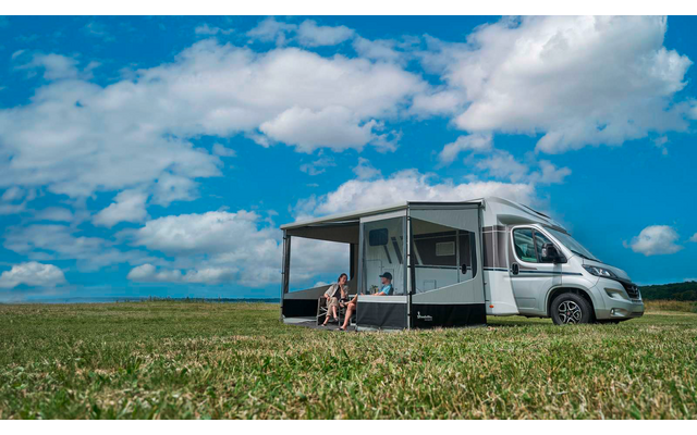 Camping-car blanc avec auvent déployé sur un champ herbeux, deux personnes assises dessous sur des chaises de camping, ciel bleu avec nuages