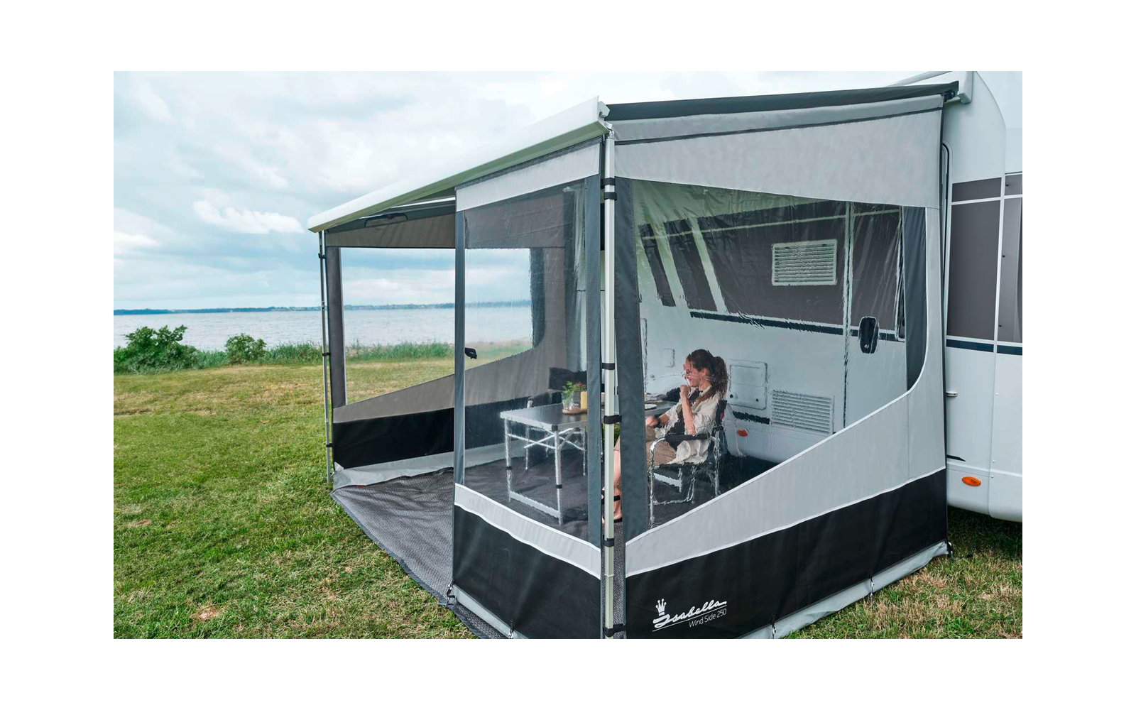 Person sitzt an einem Tisch in einem geschlossenen Vorzelt, rechts ein Wohnwagen, Wiese und See im Hintergrund, bewölkter Himmel