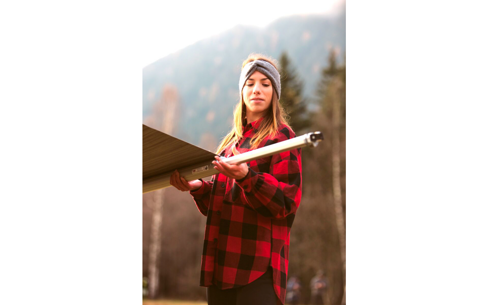 Woman in a red-and-black checkered shirt and gray headband holding a rectangular dark panel with a silver edge, blurred mountains and evergreen trees in the background