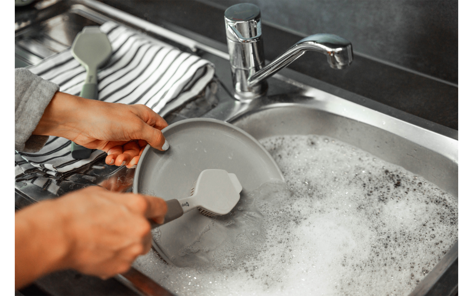 Zwei Hände halten einen grauen Teller und eine graue Spatel über einem Spülbecken mit schaumigem Seifenwasser; ein gestreiftes Geschirrtuch liegt neben dem Becken