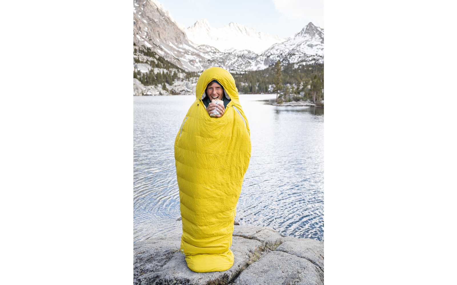 Person in gelbem Schlafsack stehend vor einem Bergsee mit schneebedeckten Bergen im Hintergrund