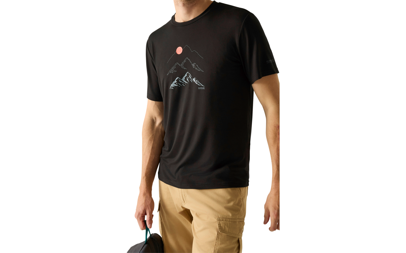 Person trägt schwarzes T-Shirt mit Bergmotiv und rosa Sonne, beige Hose, hält eine dunkle Tasche, steht vor hellem Hintergrund