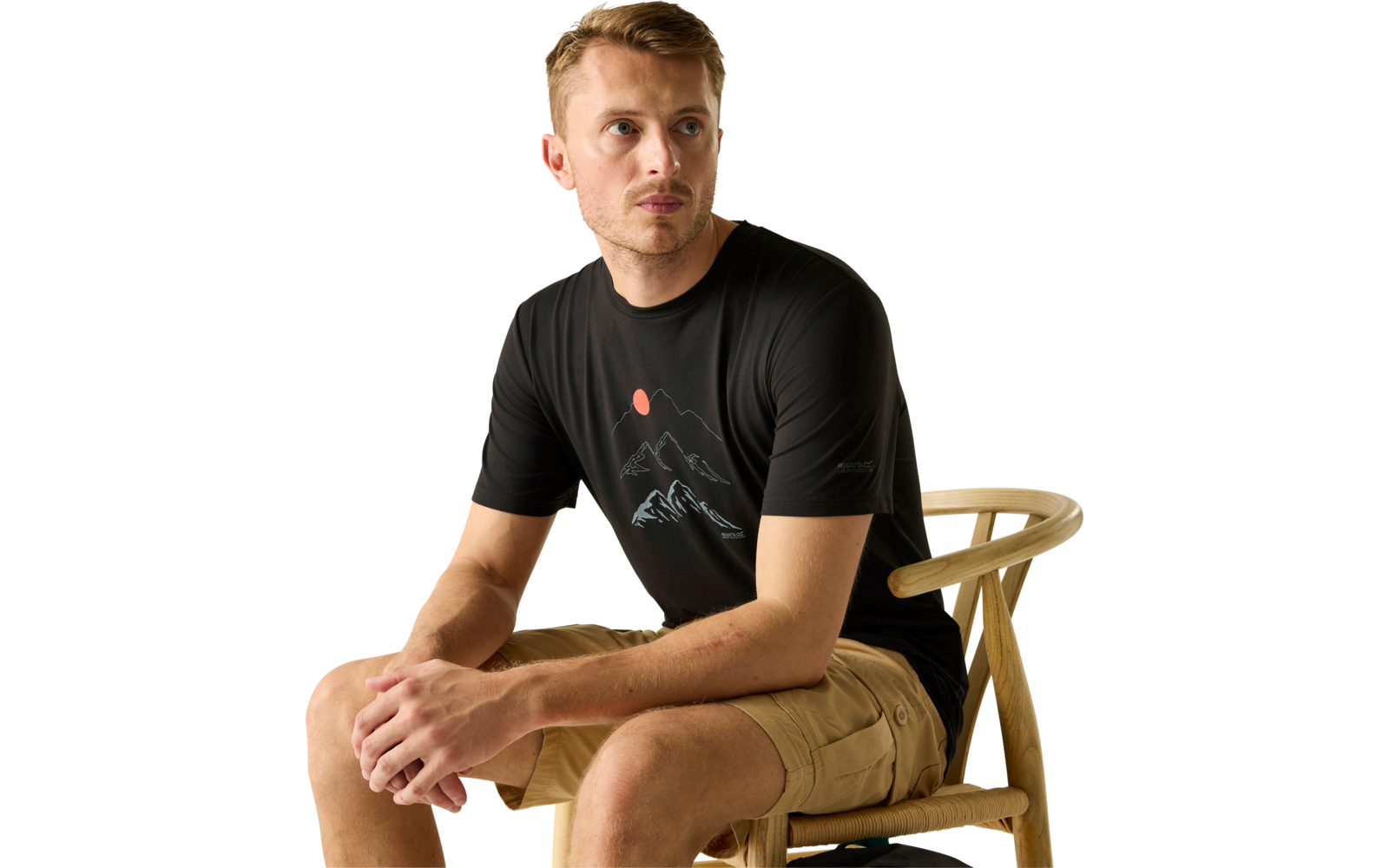 Mann sitzt seitlich auf einem Holzstuhl, trägt schwarzes T‑Shirt mit Bergmotiv und beige Shorts, schaut zur Seite vor hellem einfarbigem Hintergrund