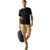 Ein Mann geht, trägt ein schwarzes T‑Shirt mit Berg- und roter Sonnen‑Grafik, beige Cargo‑Shorts, grüne Sneaker und hält einen grauen Rucksack in einer Hand vor hellem Hintergrund
