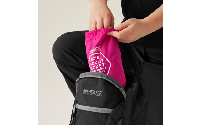 Hände schieben eine gefaltete pinke Packjacke mit dem Schriftzug 'KID'S PACK-IT JACKET' in einen schwarzen Rucksack mit Aufschrift 'REGATTA GREAT OUTDOORS'