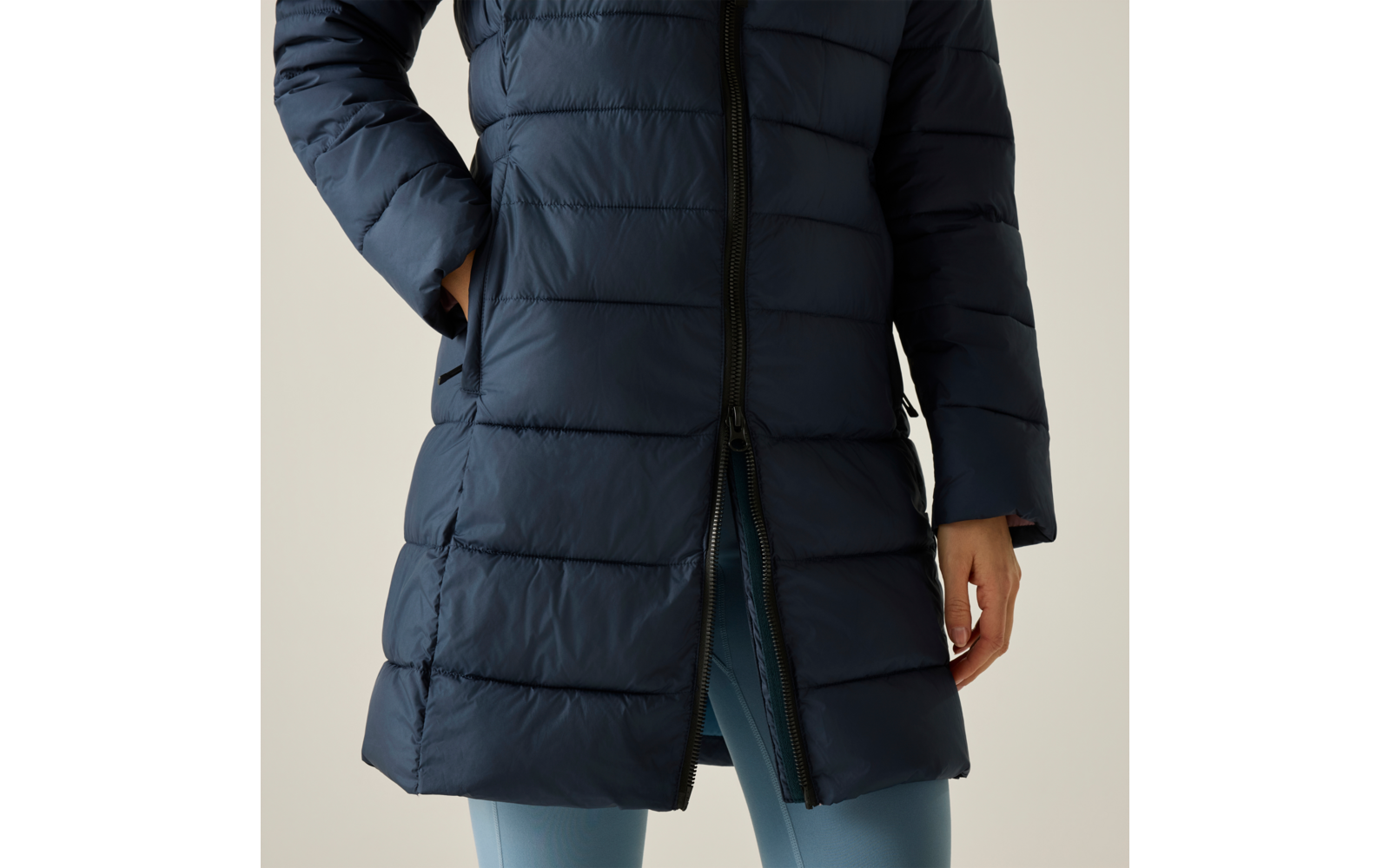 Parte centrale del corpo di una persona con un giaccone imbottito blu scuro sopra leggings azzurri, mano sinistra in tasca, mano destra visibile, cerniera inferiore aperta, sfondo chiaro