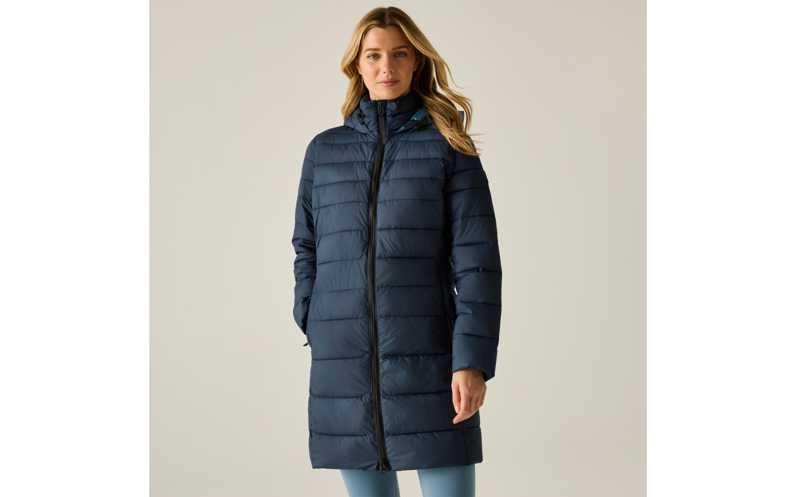 Persona con capelli biondi fino alle spalle che indossa un cappotto imbottito blu scuro fino al ginocchio con zip completa, mani nelle tasche, sfondo chiaro uniforme