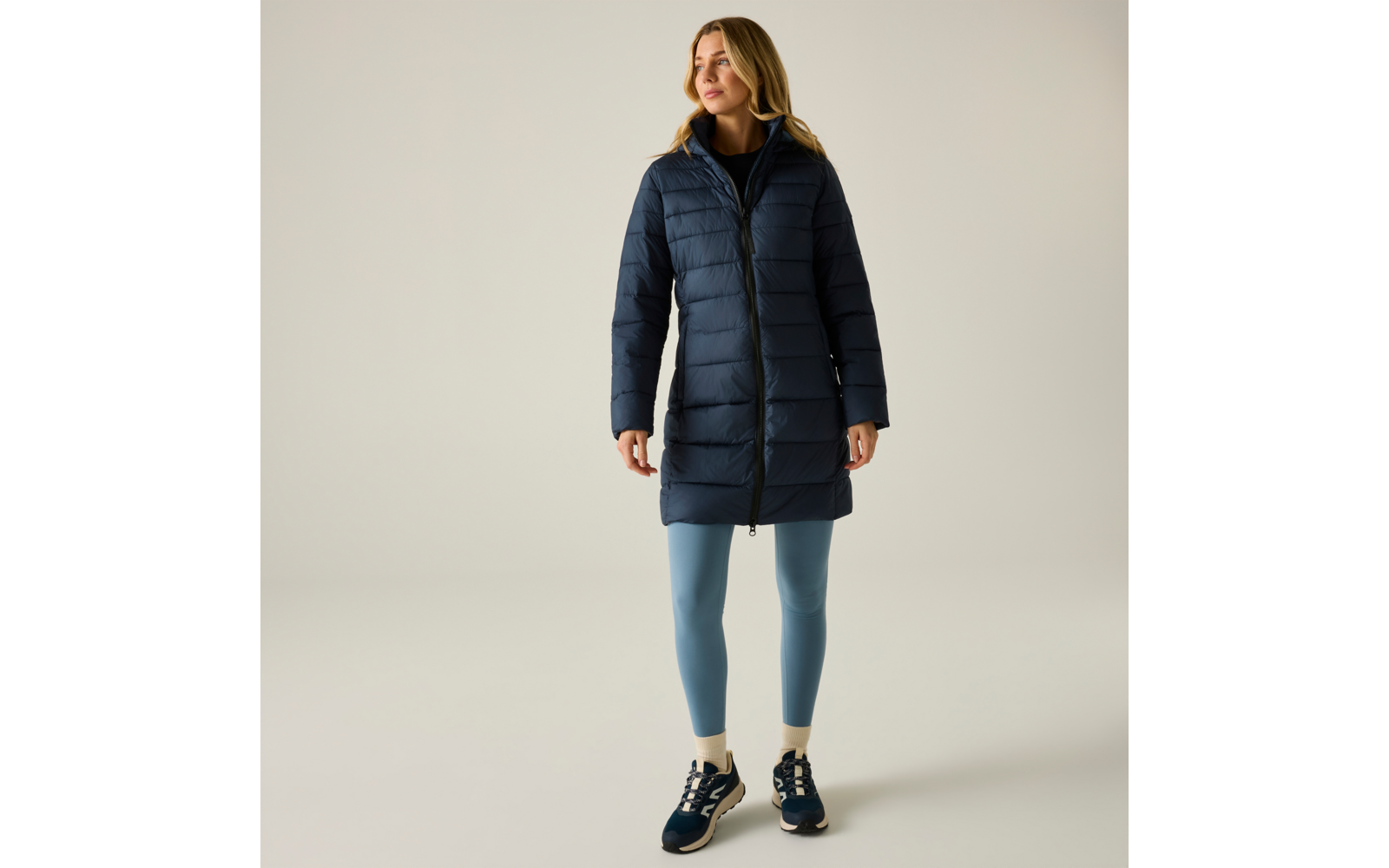 Persona in piedi su sfondo neutro, indossa un cappotto trapuntato scuro con cappuccio, leggings azzurri, sneakers scure e calzini chiari