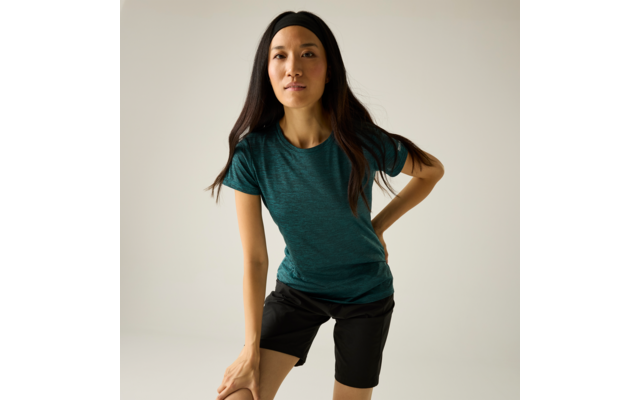 Persoon met lang donker haar, zwarte haarband, groen-blauw T-shirt en zwarte short, naar voren gebogen met één hand op een knie tegen een lichtgrijze achtergrond