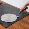 Main droite tenant une poignée noire avec une pince métallique fixée à la languette d'une plaque ronde en acier inoxydable sur une plaque de cuisson vitrocéramique noire ; plan de travail en bois et spatule en silicone visibles au bord