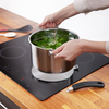 Deux mains tiennent une casserole en acier inoxydable contenant des feuilles vertes dans de l'eau sur une plaque à induction ; poignée détachée et ustensiles de cuisine sur un plan de travail en bois
