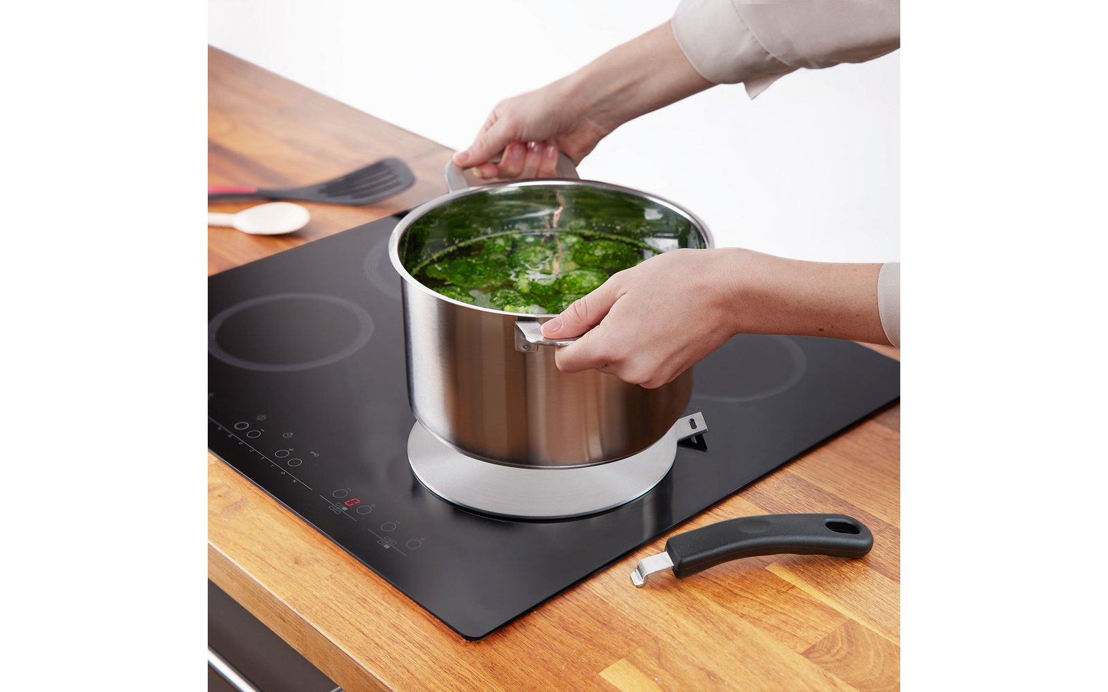 Deux mains tiennent une casserole en acier inoxydable contenant des feuilles vertes dans de l'eau sur une plaque à induction ; poignée détachée et ustensiles de cuisine sur un plan de travail en bois