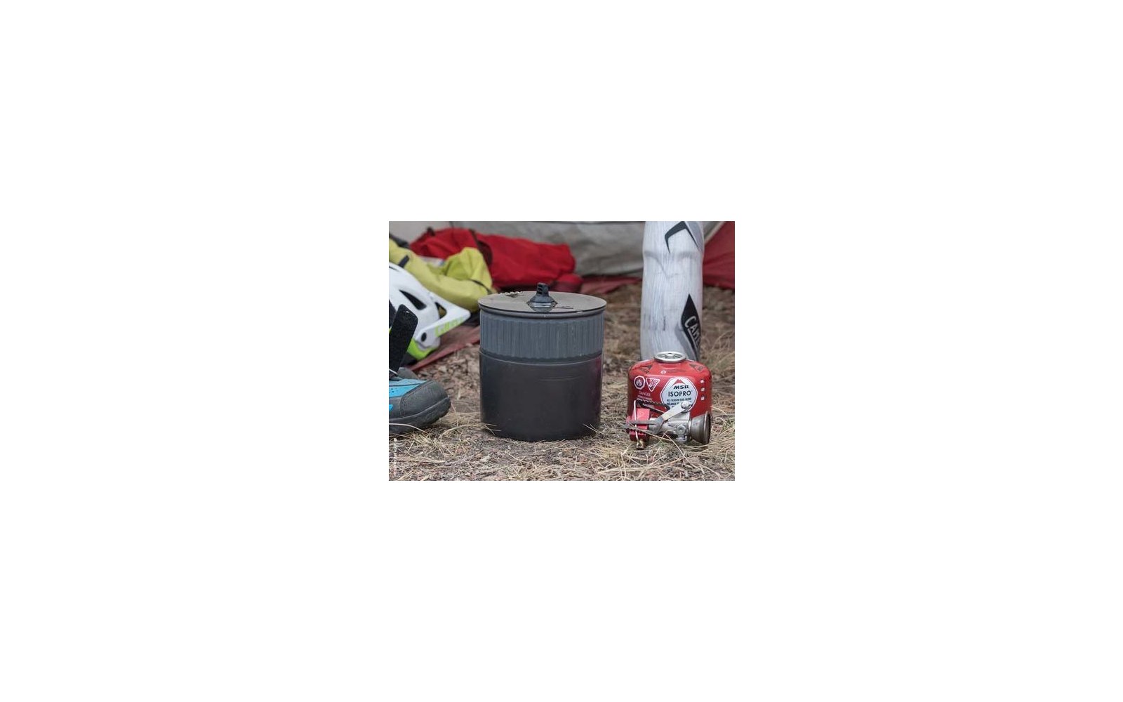 Schwarzer Campingtopf mit Deckel in der Bildmitte, rechts rote Gaskartusche mit angeschlossenem Brenner, daneben weiße Trinkflasche, links weiß-grüner Fahrradhelm und Schuh, alles auf trockenem Gras vor Zeltstoff