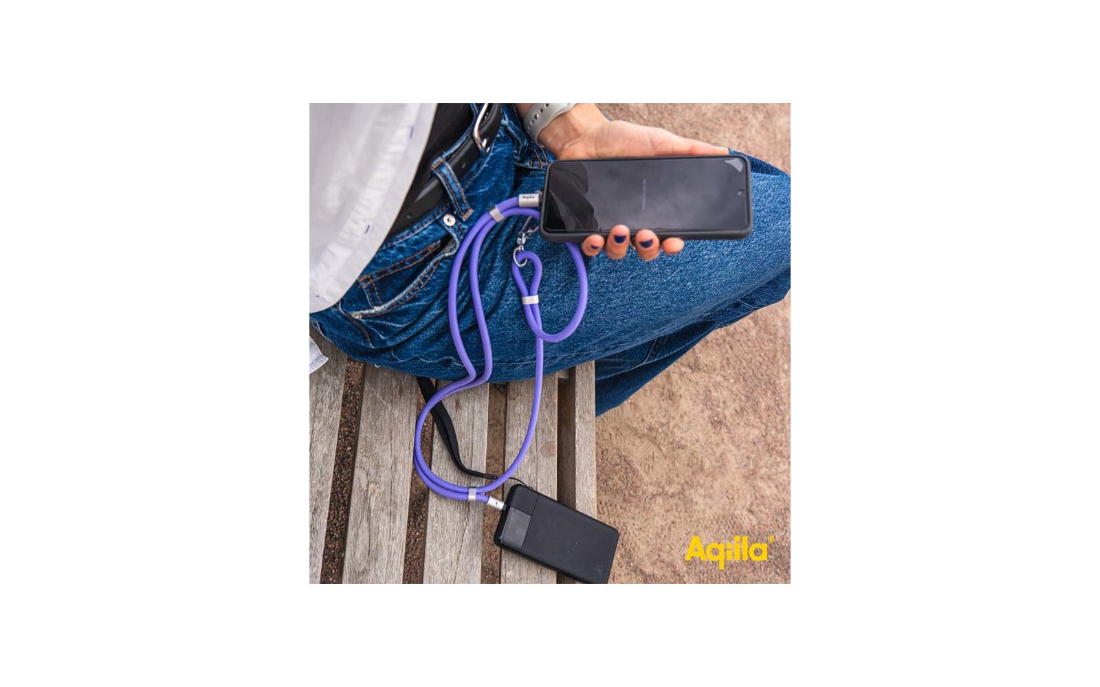 Hand hält ein Smartphone, verbunden über ein violettes Ladekabel mit einer schwarzen Powerbank auf einer Holzbank; Person trägt blaue Jeans und eine Armbanduhr; gelbes "Aqila"-Logo unten rechts