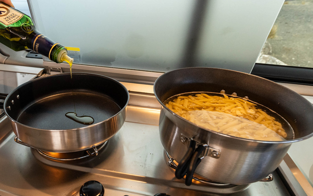 Twee pannen op een fornuis voor een mat glasraam: links een koekenpan waarin een fles olie giet; rechts een grotere pan met water en pennepasta; drie fornuisknoppen zichtbaar