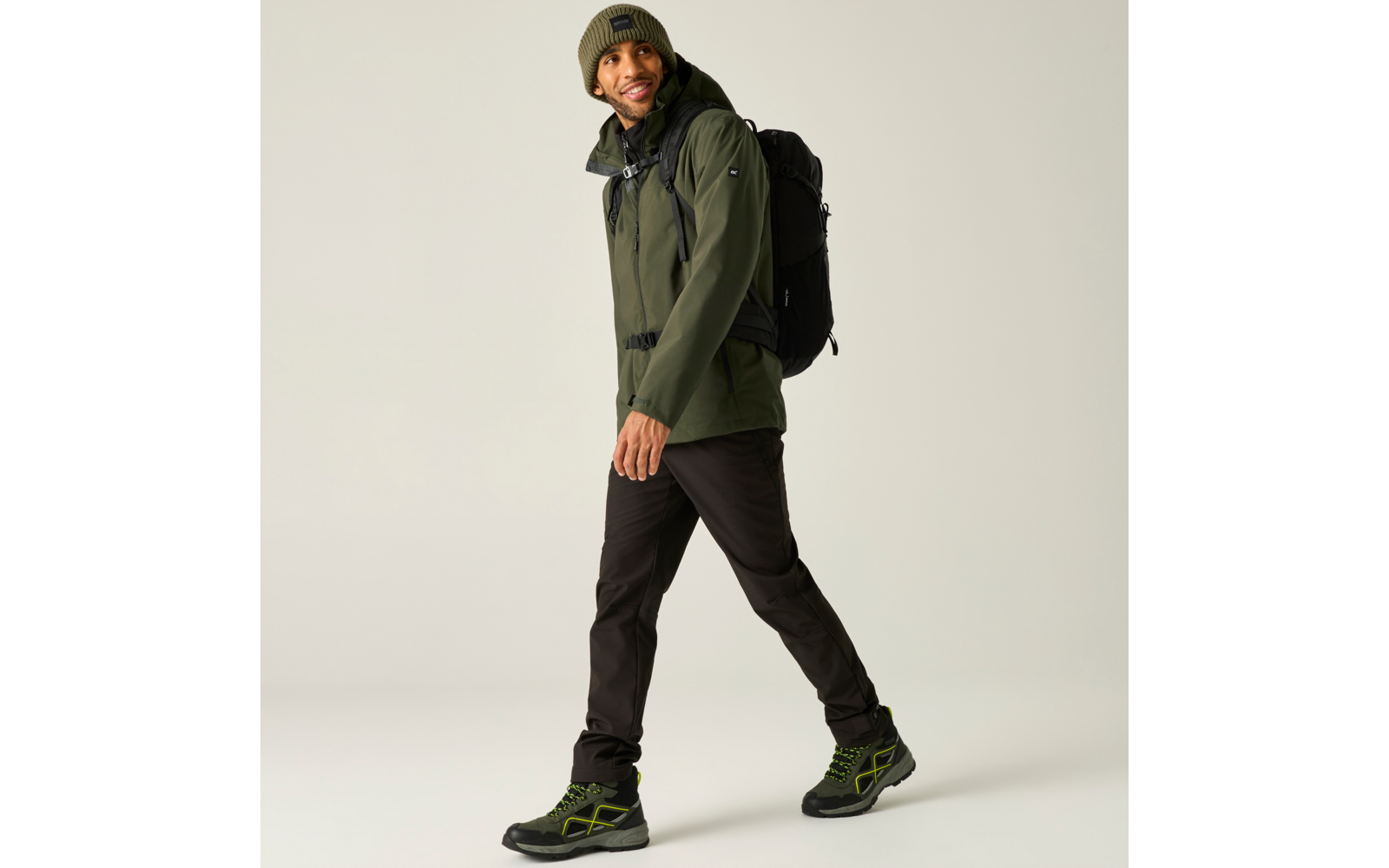 Persoon loopt, draagt een groen jack, een groene gebreide muts, een zwarte rugzak, zwarte broek en zwarte schoenen met groene details op een lichte achtergrond