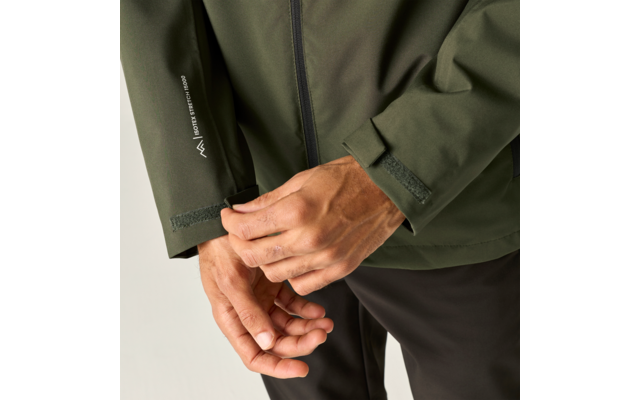 Handen die de klittenbandsluiting van de mouw van een olijfgroene jas sluiten; zichtbare ritssluiting en wit logo op de mouw; zwarte broek; lichte achtergrond