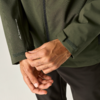 Handen die de klittenbandsluiting van de mouw van een olijfgroene jas sluiten; zichtbare ritssluiting en wit logo op de mouw; zwarte broek; lichte achtergrond