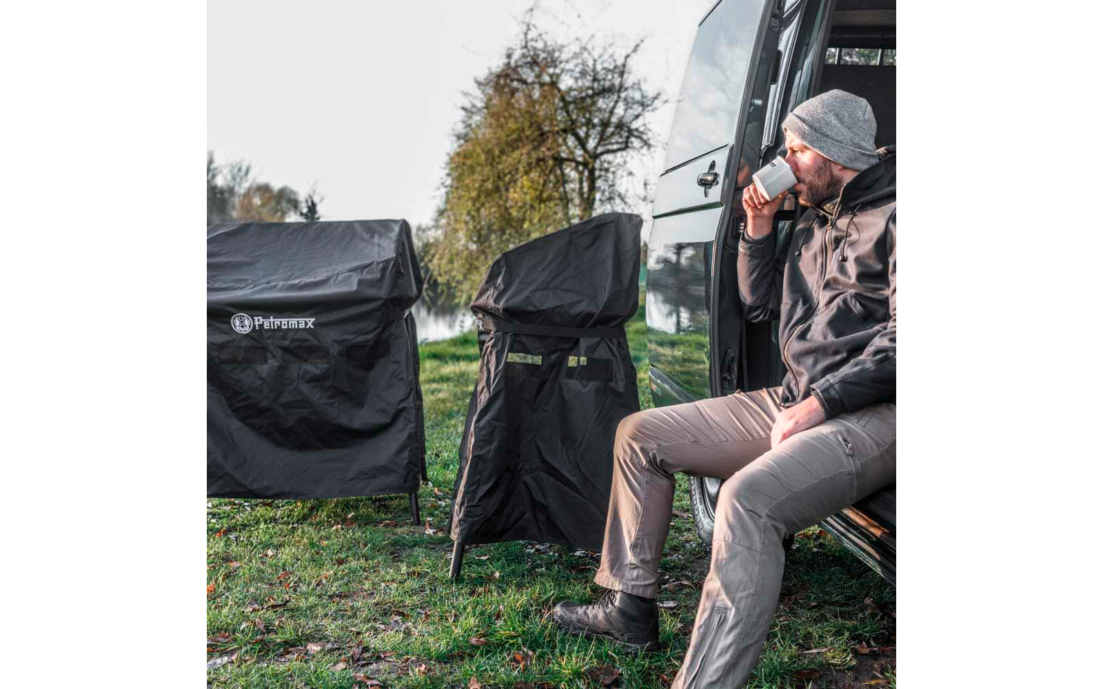 Mann sitzt in der offenen Seitentür eines Vans und trinkt aus einer Tasse; zwei mit Schutzhüllen bedeckte Stühle mit Logo neben dem Van; Gras und Bäume im Hintergrund
