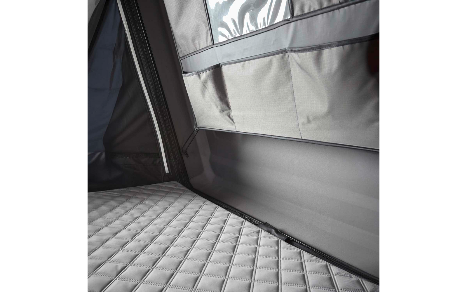 Gepolsterte, gesteppte graue Matratze in einer Ecke; darüber graue Stoffwand mit Reißverschluss, aufgesetzten Taschen und glänzendem Folieneinsatz