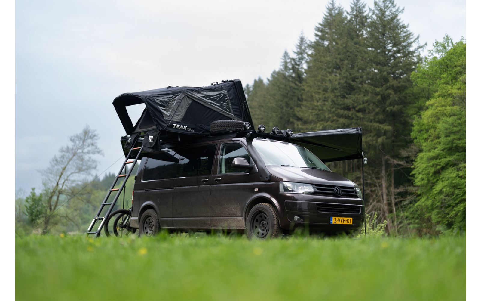 Dunkelgrauer Camper-Van mit aufgeklapptem Dachzelt, ausgeklappter Leiter und ausgefahrener Markise, auf einer Wiese vor Nadelbäumen