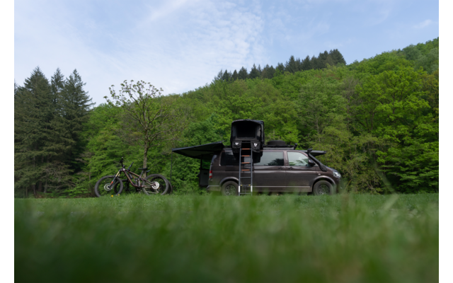 Dunkler Van auf grüner Wiese, links zwei Fahrräder, Fahrzeug mit geöffnetem Dachaufbau und Leiter; bewaldete Hügel und wolkiger Himmel im Hintergrund