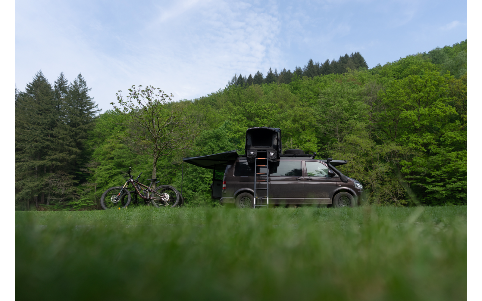Dunkler Van auf grüner Wiese, links zwei Fahrräder, Fahrzeug mit geöffnetem Dachaufbau und Leiter; bewaldete Hügel und wolkiger Himmel im Hintergrund