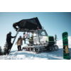 Zwei Personen neben einem cingolierten Pistenfahrzeug mit Leiter und einem aufgestellten Snowboard in schneebedeckter Landschaft mit Bäumen und blauem Himmel