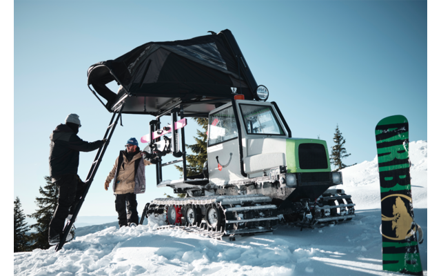 Zwei Personen neben einem cingolierten Pistenfahrzeug mit Leiter und einem aufgestellten Snowboard in schneebedeckter Landschaft mit Bäumen und blauem Himmel