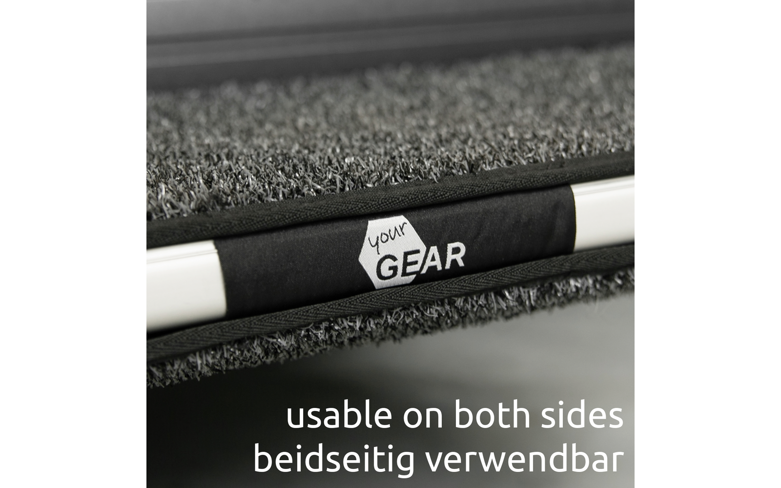 Nahaufnahme einer grauen, strukturierten Matte mit schwarzem Riemen, weißem Sechseck-Logo 'your GEAR' auf dem Riemen und einer weißen Stange; unten der Text 'usable on both sides beidseitig verwendbar'
