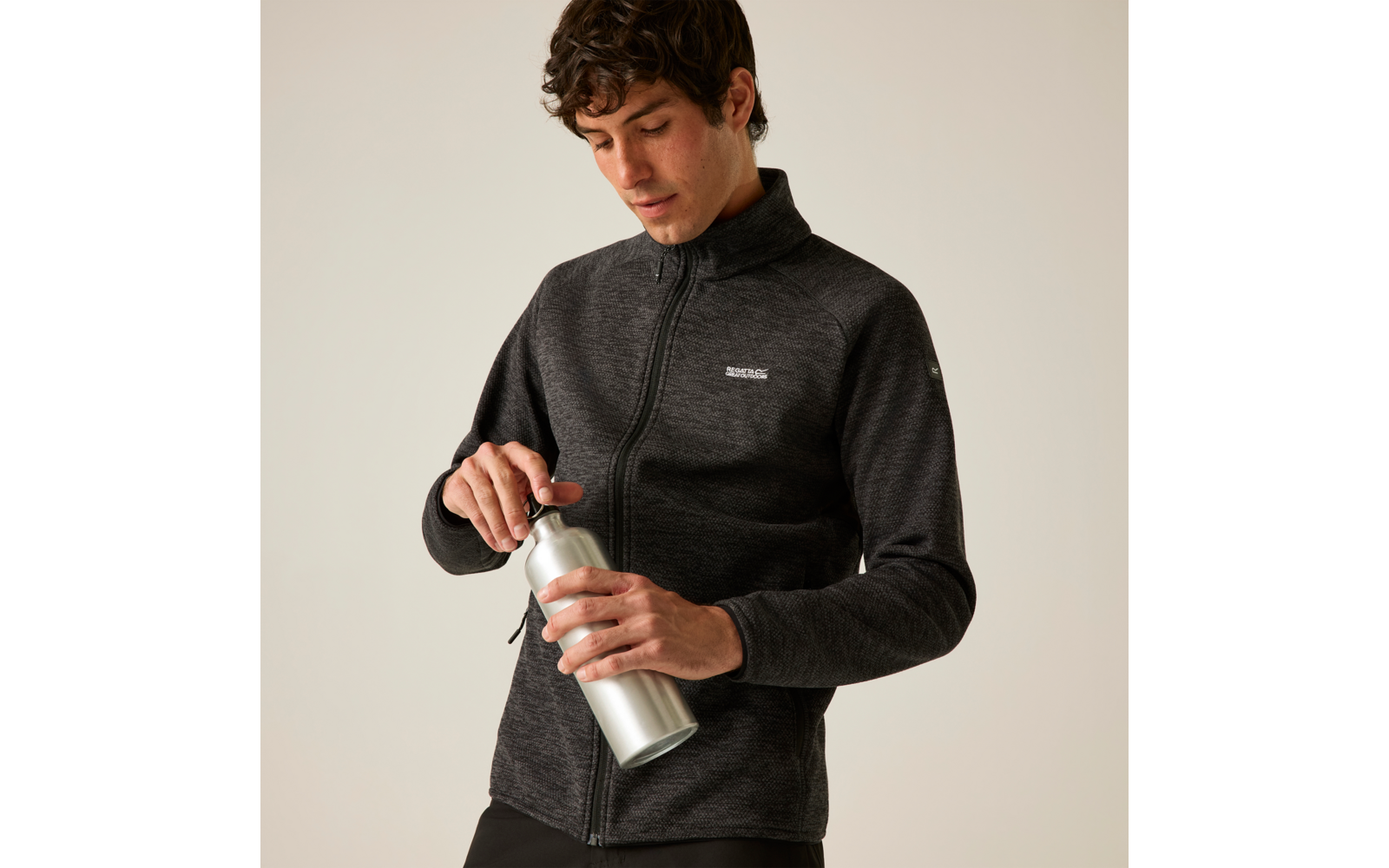 Mann in dunkelgrauer Jacke mit Reißverschluss hält eine silberne Trinkflasche und schraubt den Deckel ab, beiger Hintergrund