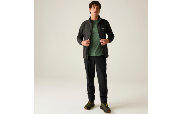 Vollkörperaufnahme einer stehenden Person vor hellem Hintergrund, die eine dunkle Reißverschlussjacke, ein grünes T‑Shirt, schwarze Hose und dunkle Wanderschuhe trägt