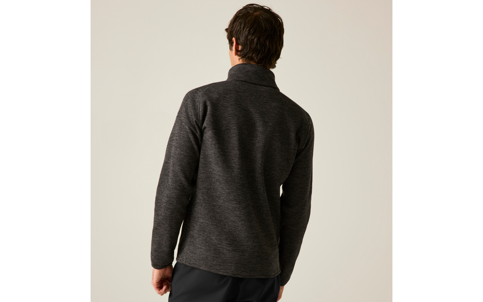 Rückenansicht einer Person mit kurzen dunklen Haaren, die einen dunkelgrauen Pullover mit Stehkragen und schwarze Hosen vor hellem Hintergrund trägt