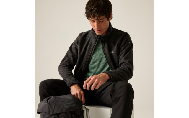 Person sitzt auf einem Hocker, trägt dunkle Fleecejacke, grünes T‑Shirt und schwarze Hose, greift zu einem Rucksack neben sich