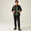 Vollkörperaufnahme einer stehenden Person vor hellem Hintergrund, die eine dunkle Reißverschlussjacke, ein grünes T‑Shirt, schwarze Hose und dunkle Wanderschuhe trägt