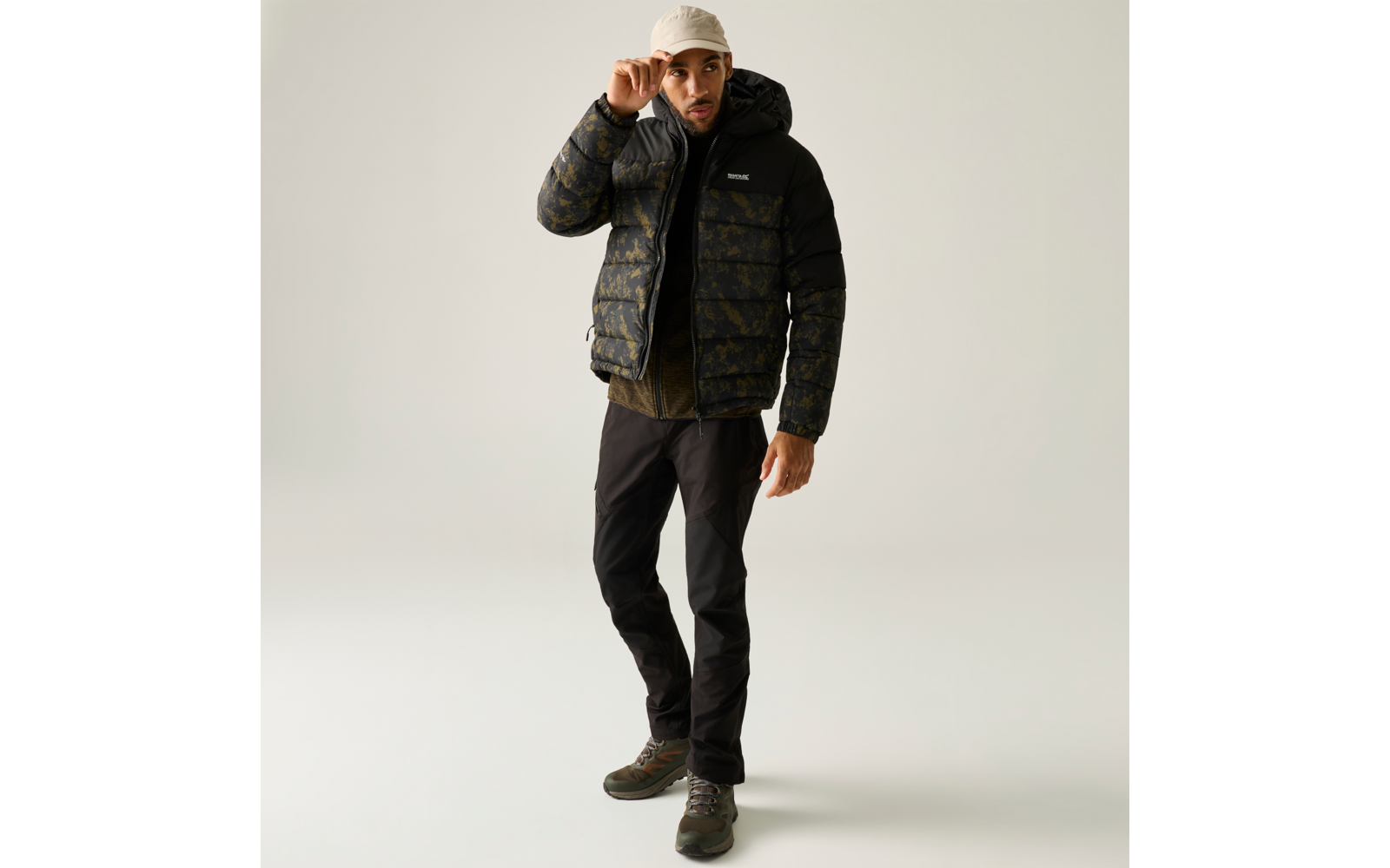 Mann steht vor neutralem Hintergrund, trägt beige Baseballkappe, schwarze gesteppte Camouflage-Jacke mit Kapuze, dunkle Hosen und grüne Schuhe; er berührt mit der rechten Hand die Kappe