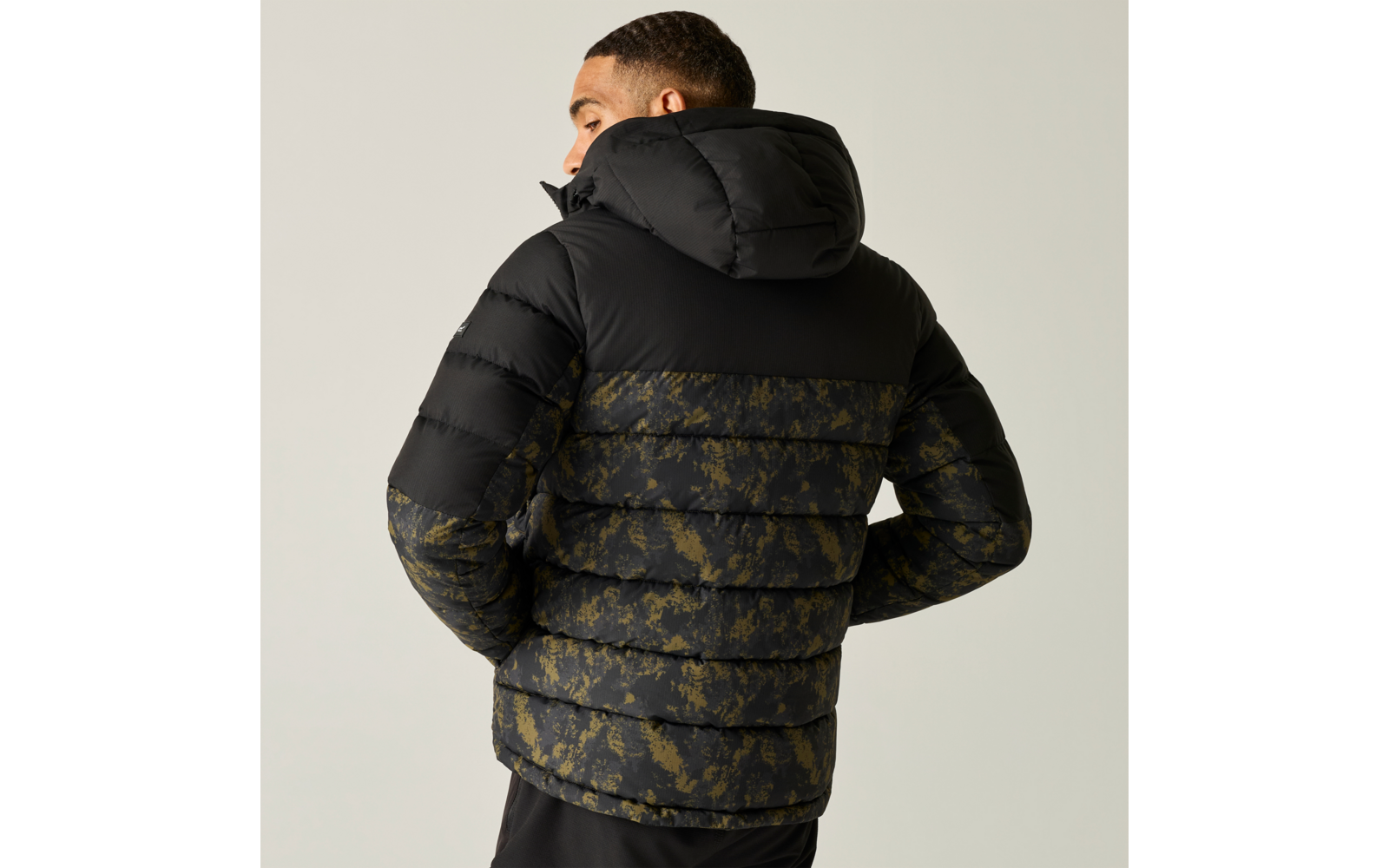 Person von hinten, trägt schwarze Steppjacke mit Kapuze und olivgrünem Camouflagemuster auf unterem Rücken und Ärmeln, Hände in den Taschen, heller Hintergrund