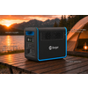 Schwarz-blaues tragbares Powerstation-Gerät mit Display, mehreren Steckdosen und Lüftungsgitter auf einem Holztisch; im Hintergrund Zelt, Lampe, Bergkette und Sonnenuntergang