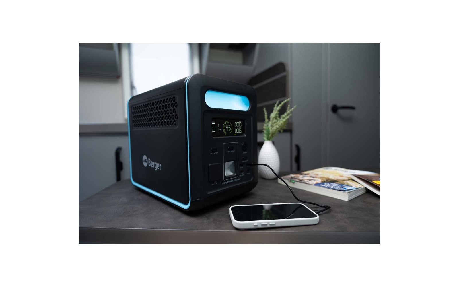 Schwarzes würfelförmiges Gerät mit blauem Leuchtrand und Display auf einem Tisch; daneben liegt ein mit einem Kabel verbundenes Smartphone, eine weiße Vase mit Pflanze und Zeitschriften