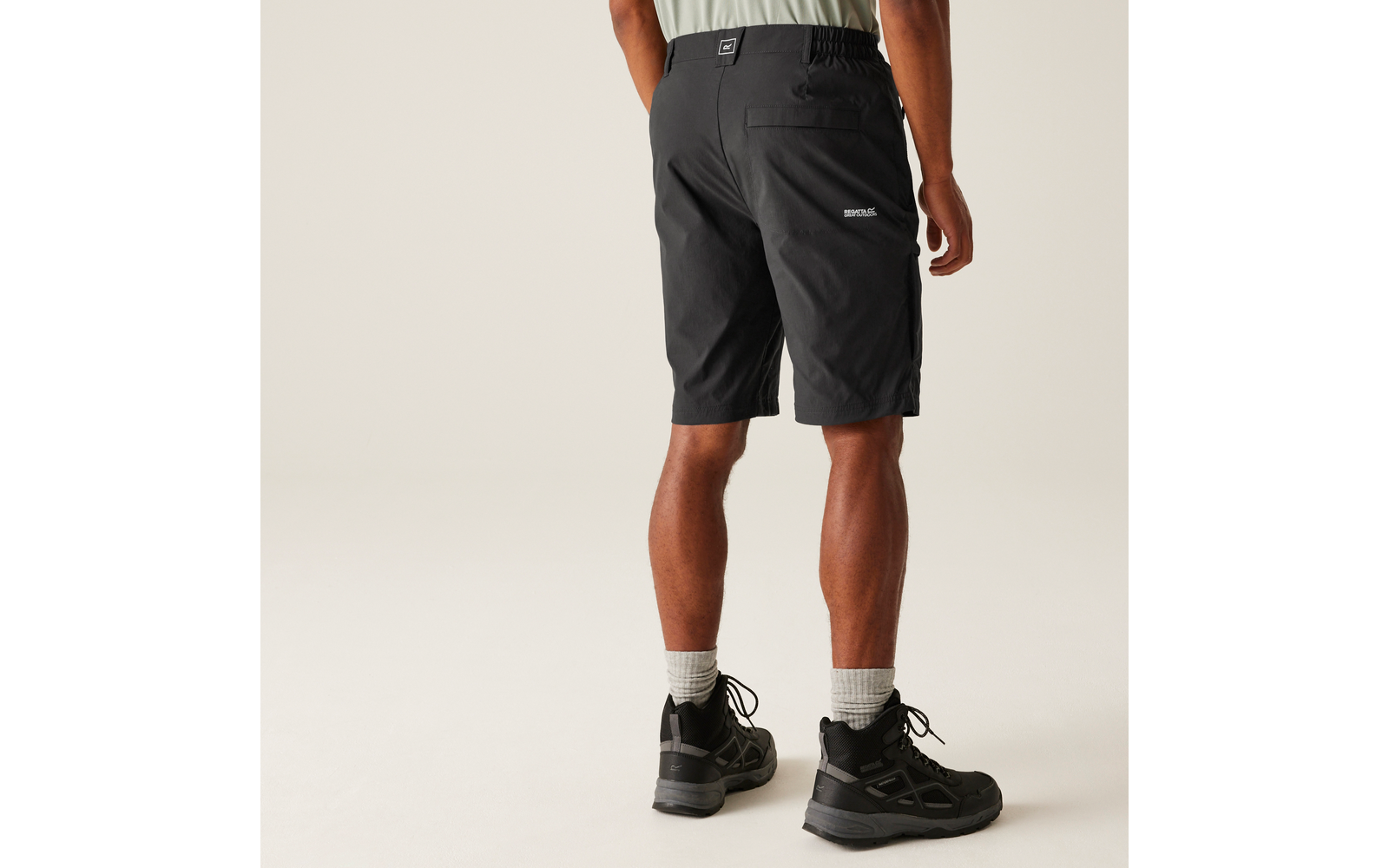 Person von hinten, knielange schwarze Shorts, helles T-Shirtansatz sichtbar, weiße Socken, schwarze Schnürschuhe, heller neutraler Hintergrund