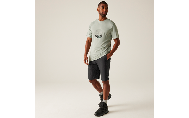 Mann steht vor hellem Hintergrund, trägt hellgrünes T‑Shirt mit rundem Print, dunkle knielange Shorts und schwarze Wanderschuhe; eine Hand in der Hosentasche